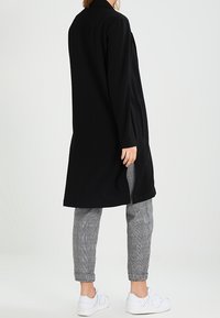 Svart lång kappa med rakt snitt och sidofickor, bärs över grå rutiga byxor. Har långa ärmar och krage.