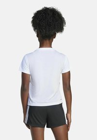 Persona con pelo rizado de pie de espaldas, vistiendo una camiseta blanca de manga corta lisa y pantalones cortos deportivos negros con rayas blancas en los lados.
