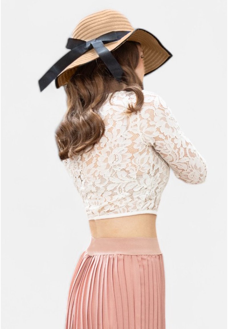 Femme aux cheveux ondulés portant un chapeau beige à large bord avec un ruban noir, un crop top en dentelle blanche à manches longues et une jupe plissée rose, vue de dos.