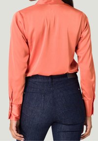 Chemise en satin corail à manches longues avec poignets boutonnés, portée avec un jean en denim foncé présentant deux poches arrière et un léger effet délavé.