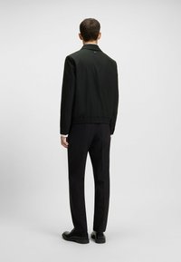Black lightweight jacket with elasticated hem and collar, paired with tailored black trousers and black shoes. Clean, minimalist design.