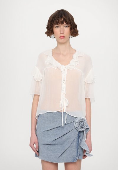 Blouse blanche et transparente avec des manches à volants et un détail noué à l'avant, associée à une jupe en denim ornée d'un accent floral à la taille.