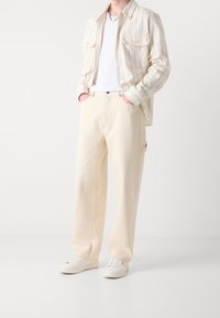 Pantalon large beige clair en coton texturé, associé à un t-shirt blanc et une chemise rayée de couleur crème. Des baskets blanches complètent la tenue.