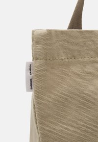 Beige lerretsekk med en teksturert overflate, med en sidemerke som har "SAMSØE" i svart, og et enkelt, strukturert design.