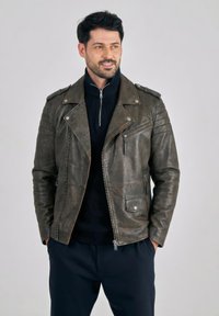 Man draagt een donker groene leren motorjack over een zwart vest met ritssluiting, met handen in de zakken, kijkt opzij tegen een effen achtergrond.