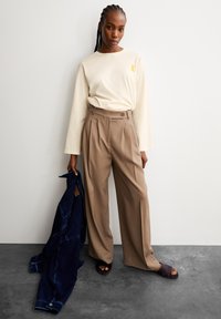 Langärmliges cremefarbenes Hemd mit einem gelben Band, hoch taillierte beige Hose, hält eine dunkelblaue Jeansjacke und trägt dunkle Sandalen.