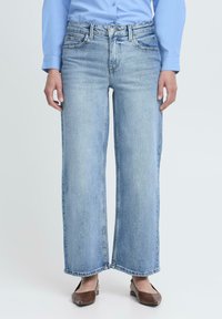 Šviesiai mėlynos, plačios kojos džinsai su aukštu liemeniu, priekinėmis kišenėmis ir detalizuotomis siūlėmis. Nešioti su rudais butais plokščiu padu, atrodantys laisvai.