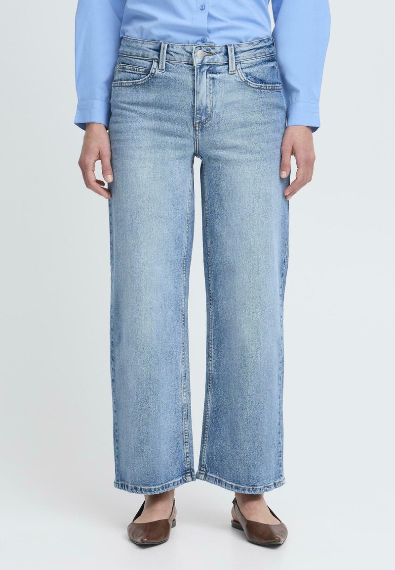 Šviesiai mėlynos, plačios kojos džinsai su aukštu liemeniu, priekinėmis kišenėmis ir detalizuotomis siūlėmis. Nešioti su rudais butais plokščiu padu, atrodantys laisvai.