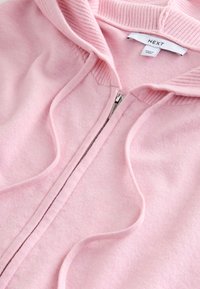Jasnoróżowa bluza na zamek wykonana z miękkiego materiału, z prążkowanym kołnierzem i troczkami, z widocznym srebrnym zamkiem oraz metką marki.