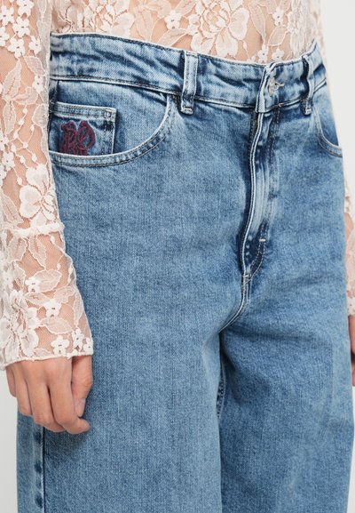 Jeans de mezclilla azul claro con un corte relajado, que presentan un cierre delantero, presillas para cinturón y un diseño bordado rojo en el bolsillo trasero.
