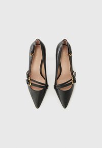 Talons hauts en cuir noir avec un bout pointu, arborant des straps superposés et des accents en métal doré. L'intérieur est beige.