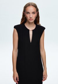 Vestido negro sin mangas con escote en V y un acento dorado en el cuello. Presenta pliegues verticales y una silueta ajustada. Textura de tela suave.