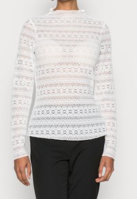 Vit långärmad spets-topp med blommiga och geometriska mönster, hög krage och figurnära siluett, tillsammans med svarta byxor.