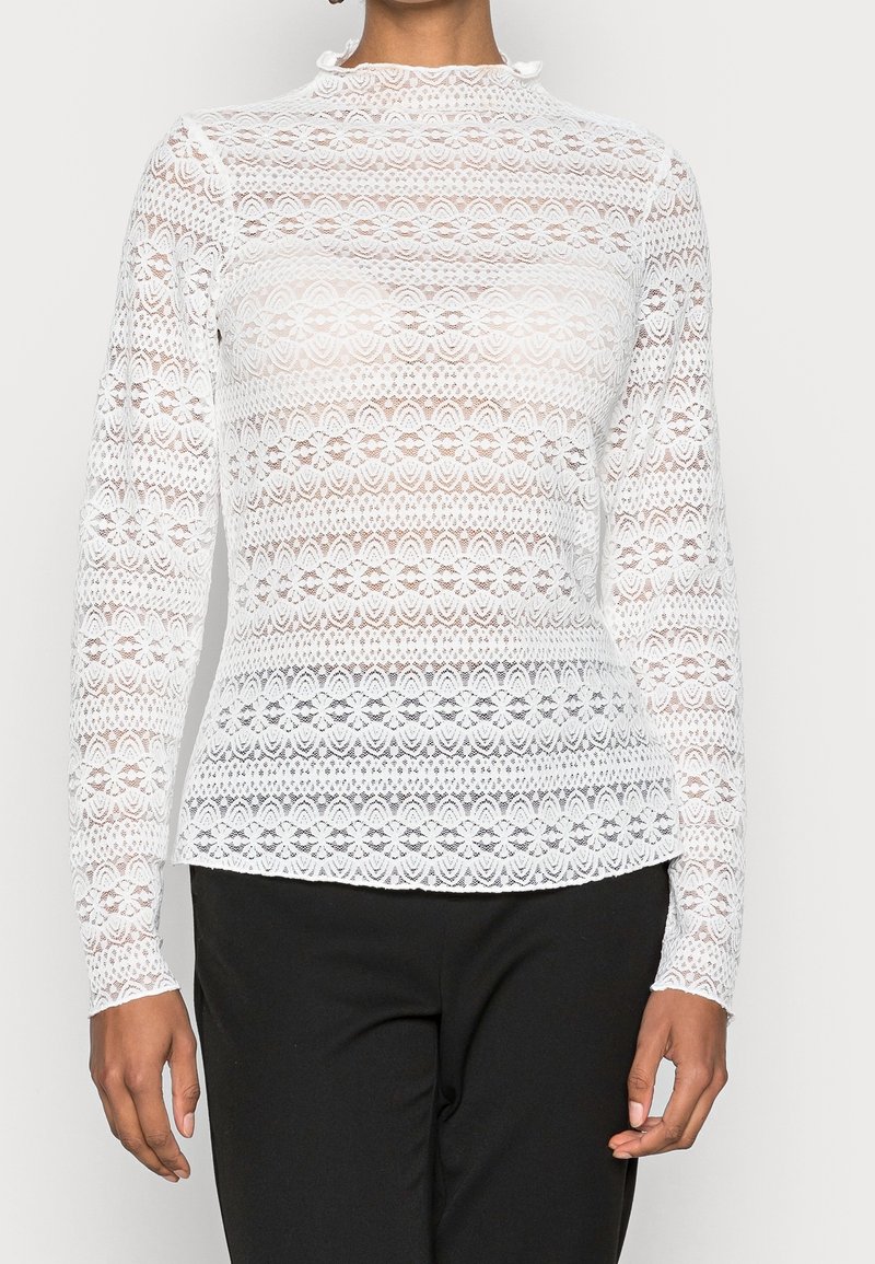 Vit långärmad spets-topp med blommiga och geometriska mönster, hög krage och figurnära siluett, tillsammans med svarta byxor.