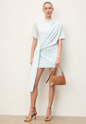 Woman wearing a light blue ruched mini dress with draped detail, holding a brown suede handbag, and wearing woven high-heeled sandals.