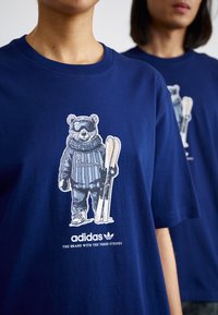 Tamsiai mėlynas medvilninis marškinėliai su grafiniu spausdiniu, vaizduojančiu lokį slidininko aprangoje, laikantį slidės, o po dizainu yra balti adidas logotipai.