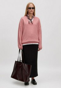 Overmaatse roze trui met v-hals, zwarte maxi rok, glanzende bordeauxrode handtas, zwarte instappers en een geruite sjaal om de hals.
