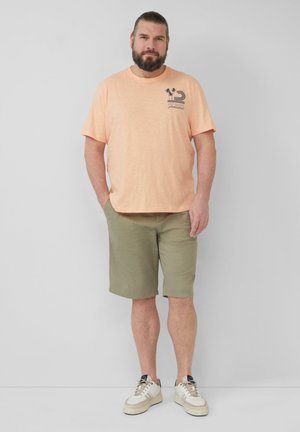 Hellorange T-Shirt mit kurzen Ärmeln und navyblauem Grafikdruck, kombiniert mit grünen Shorts und weißen Sneakern mit beigen Akzenten.