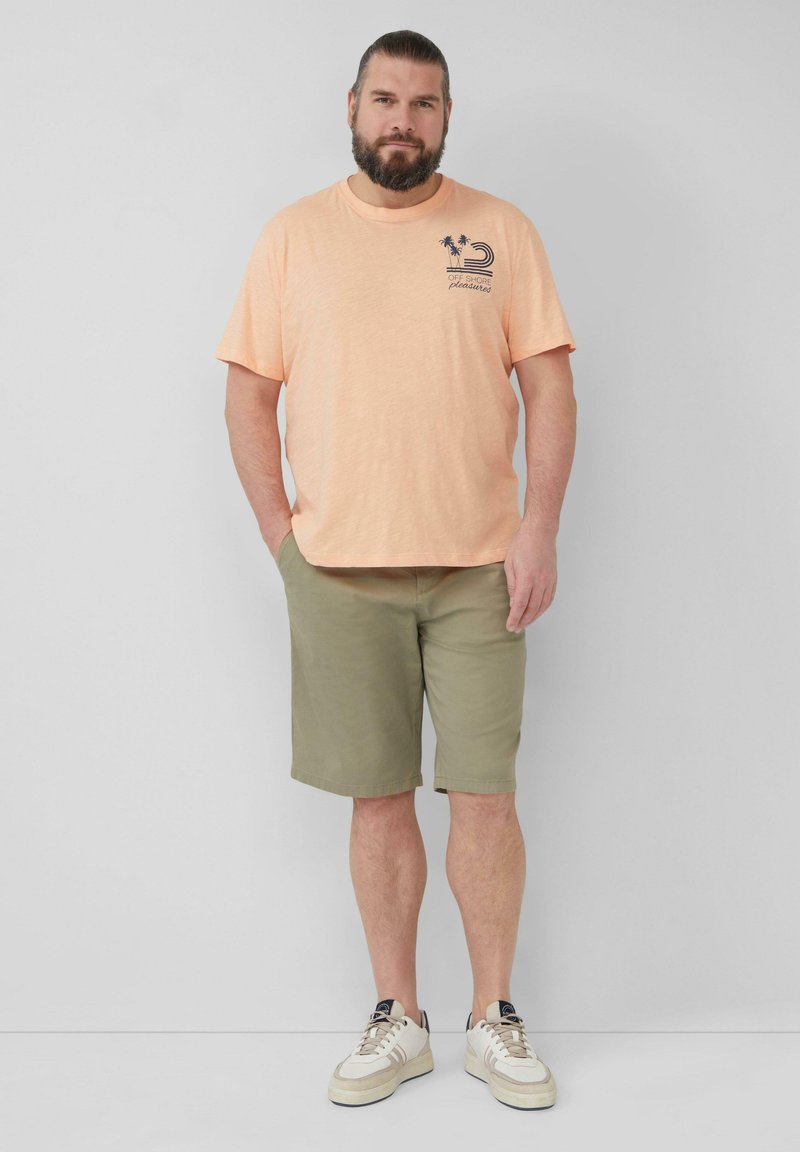 Hellorange T-Shirt mit kurzen Ärmeln und navyblauem Grafikdruck, kombiniert mit grünen Shorts und weißen Sneakern mit beigen Akzenten.