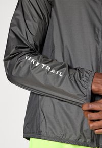 Letvægts grå løbejakke med elastiske manchetter, der har "NIKE TRAIL" logo i hvidt på ærmet og en tekstureret stoffinish.