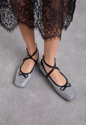 Pieds portant des ballerines tissées argentées avec des brides noires et de petits nœuds, associées à une jupe en dentelle noire sur un sol gris.