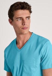 Korte mouw, turquoise t-shirt gemaakt van gladde katoenen stof. V-hals ontwerp met een enkele voorzak. Eenvoudige en schone esthetiek.