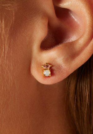Boucle d'oreille en or avec une pierre blanche centrale et un accent en gemme rose au-dessus, sur fond d'un gros plan d'une oreille.