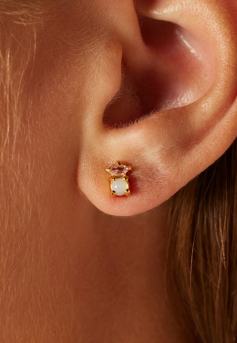 Goldene Steckerohrringe mit einem zentralen weißen Stein und einem rosa Schmuckakzent darüber, vor einem Nahaufnahme eines Ohrs.