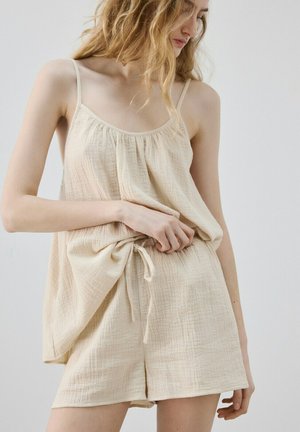 Žena koja podešava labavu, krem boje, majicu bez rukava i odgovarajuće kratke hlače od teksturiranog laganog tkanja.