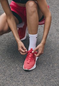 Røde sportsko med strukturerte overflater, hvit såle og flate lisser. Personen knytter lisser mens han har på hvite sokker.