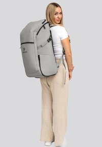 Sac à dos gris avec des accents et des fermetures éclair noires, comportant des bretelles réglables et un logo, porté sur le dos sur un fond neutre.