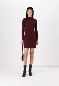 Vestido mini em burdeos com detalhes em relevo, mangas longas, gola alta e bainha desfiada. Combinado com botas pretas até ao tornozelo e uma pequena bolsa preta.