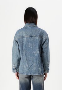Denim jakna s sproščenim krojem, ki ima bele kovinske žebljičke v vzorcu zvezdne eksplozije na hrbtu in klasično obliko ovratnika.