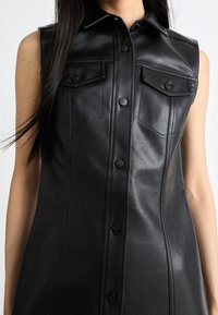 Zwarte leren mouwloze jurk met een spits kraag, knoopsluiting aan de voorkant en twee borstzakken. Gladde textuur en getailleerde pasvorm.