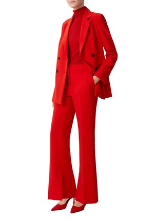Femme portant un blazer rouge cintré, un pantalon évasé taille haute assorti, un col roulé rouge et des escarpins rouges à bout pointu, debout avec une main dans la poche.