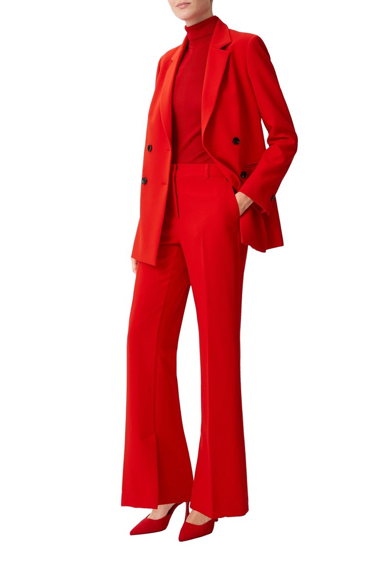 Femme portant un blazer rouge cintré, un pantalon évasé taille haute assorti, un col roulé rouge et des escarpins rouges à bout pointu, debout avec une main dans la poche.