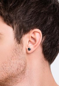 Orecchino a forma di brillantino nero con una montatura in argento, indossato nel lobo. I capelli sono corti e castani, con una leggera peluria visibile sulla linea della mascella.