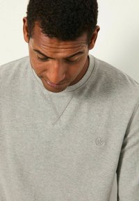 Grauer Strickpullover mit strukturiertem Finish, mit Rundhalsausschnitt und kleinem Logo auf der Brust. Der Stoff wirkt weich und gemütlich.