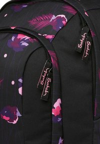 Sac à dos noir avec un motif floral rose et violet, doté de deux compartiments zippés avec des tirettes noires portant l'inscription "Satch" en texte rose.