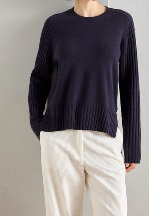 Femme portant un pull côtelé bleu marine foncé et un pantalon en velours côtelé écru, debout avec une main dans la poche devant un fond uni.