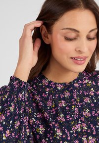 Blusa floral con fondo azul marino oscuro, con flores rosas, blancas y verdes. Mangas largas abullonadas con puños fruncidos y detalle de botones.