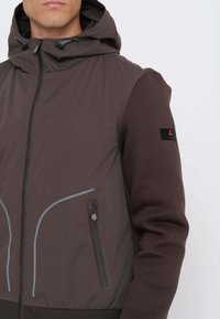 Uomo che indossa una giacca con cappuccio di colore marrone scuro con cerniera e tasca laterale con cerniera, caratterizzata da una rifinitura riflettente e una toppa nera sulla manica sinistra.