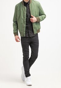 Zielona kurtka bombowca z prążkowanymi ściągaczami przy mankietach i kołnierzu, noszona na czarnej koszuli na guziki oraz zniszczonych czarnych dżinsach. Białe tenisówki dopełniają całość.