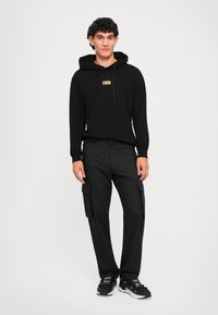 Jovem de pé, virado para a frente, vestindo um hoodie preto com um emblema, calças de carga pretas e ténis pretos com solas brancas.