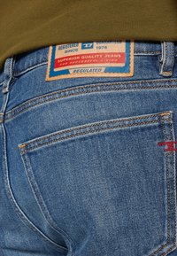 Modré džíny s kapsou vzadu a hnědou a modrou etiketou s nápisem "Superior Quality Jeans" a osoba oblečená v olivově zelené košili.