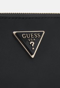 Mustapahka lompakko, jossa on kolmiomainen kultakoristeinen logo, jossa lukee "GUESS USA" ja kysymysmerkki. Vetoketjuyksityiskohdat ylhäällä.