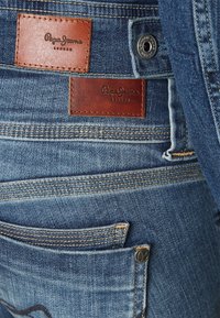 Modré džínové kalhoty z denimu s koženými štítky s nápisem "Pepe Jeans London", kontrastním prošíváním a klasickým pětipytlovým designem.