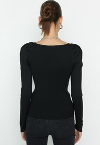 Zwarte geribbelde top met lange mouwen, een vierkante halslijn en opgeblazen schouderdetails. Zit nauwsluitend om het lichaam. Gecombineerd met zichtbare zwarte jeans.