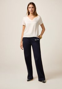 Blusa de seda con escote en V y mangas cortas de color crema, combinada con pantalones de pierna ancha en color marino que presentan bordado "Sport Chic". Unos tacones negros con tachuelas completan el conjunto.
