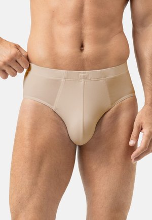 Mann in beiger Unterwäsche dehnt den Bund auf der linken Seite und zeigt Passform und Elastizität vor einem schlichten hellen Hintergrund.